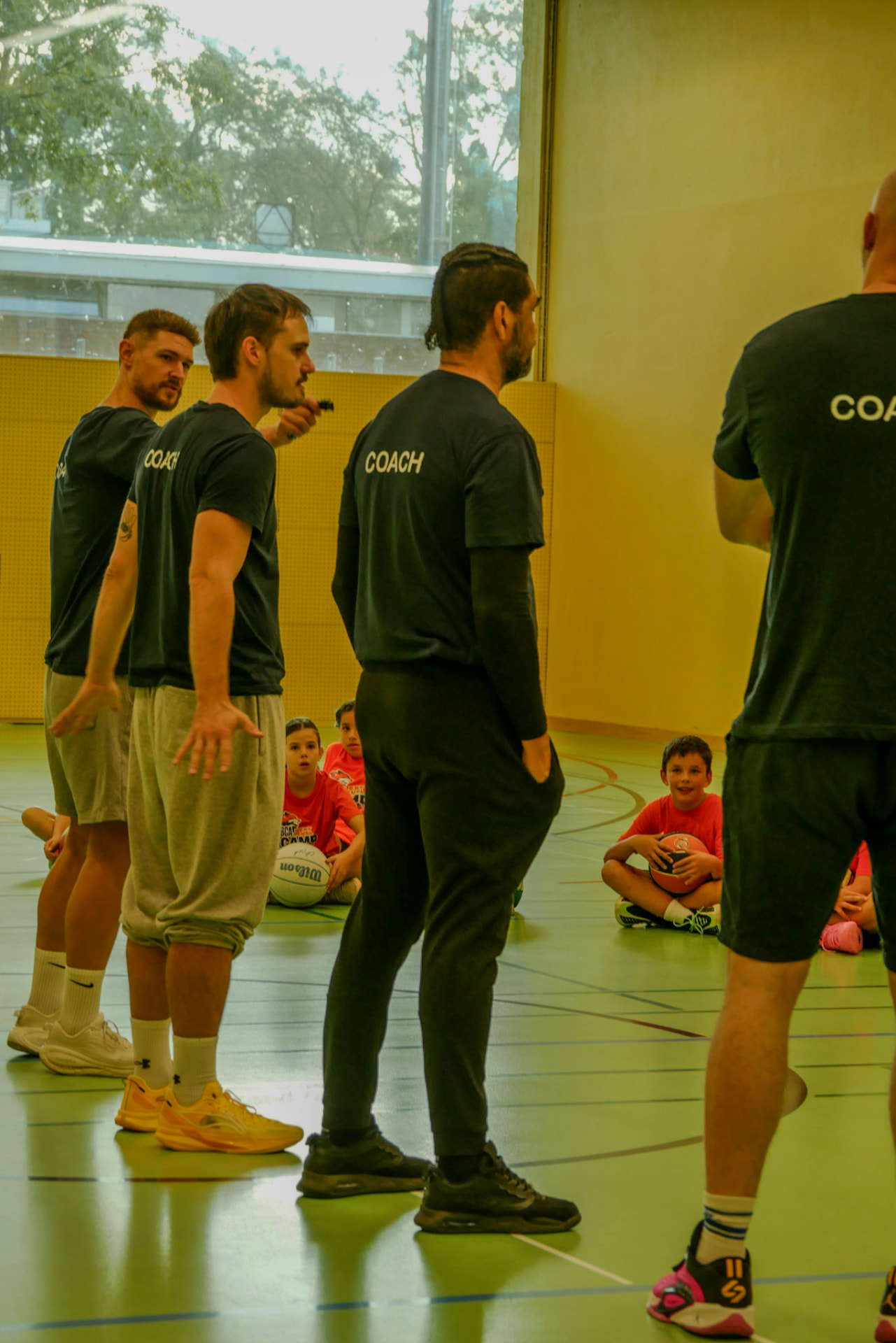 Trainer in dunklen Camp-Shirts erklären einer Gruppe sitzender Camper die nächste Übung